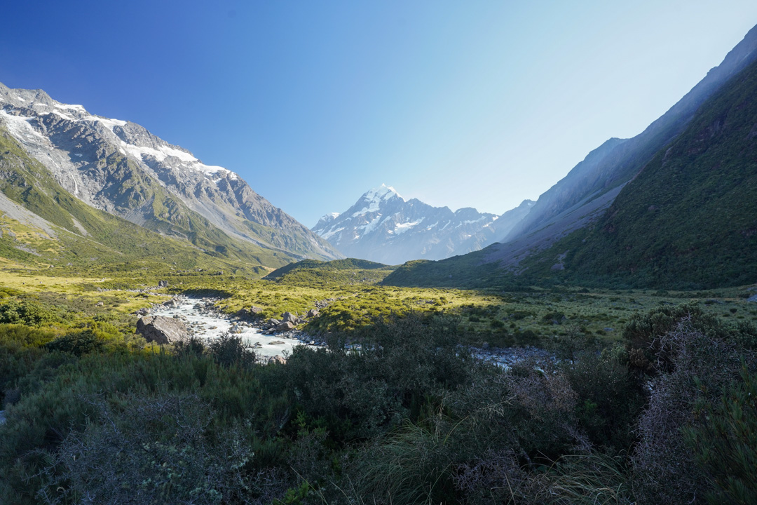 A glacial river winding through the green Hooker Valley toward snow-covered Aoraki / Mount Cook under a clear sky.